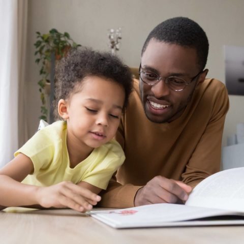 African Father And Son Lying On Warm Floor Reading Book