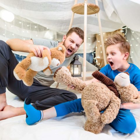 Excited Father And Son Sitting In Blanket Fort And Playing With