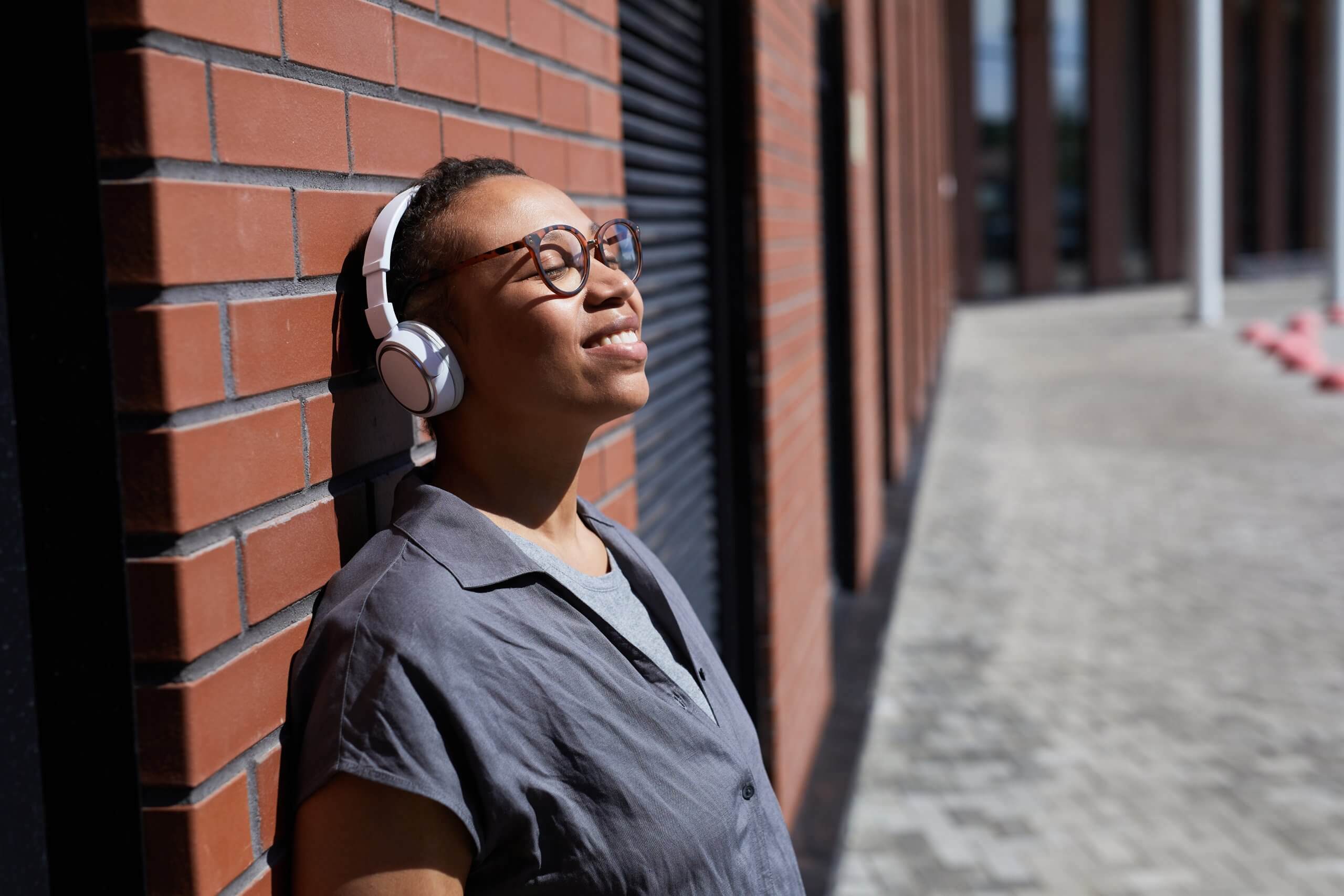 African Happy Woman In Wireless Headphones Leaning On The Brick